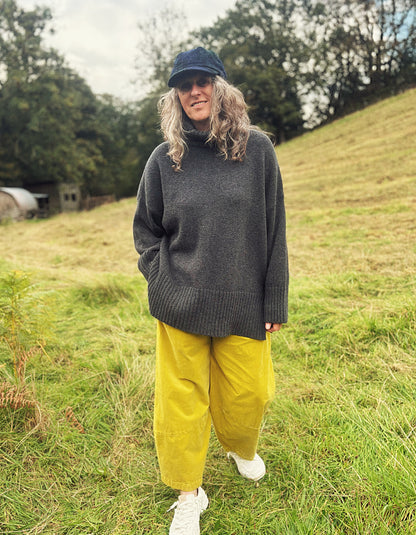 A woman standing outdoors on a grassy hill wearing an oversized dark gray turtleneck sweater, mustard yellow pants, white sneakers, and a dark blue cap.