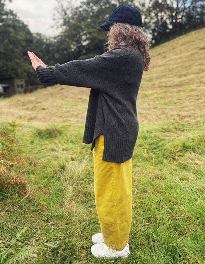 A person standing outdoors in a grassy field, wearing a dark charcoal knit cardigan, mustard yellow corduroy trousers, a dark navy cap, and white sneakers.