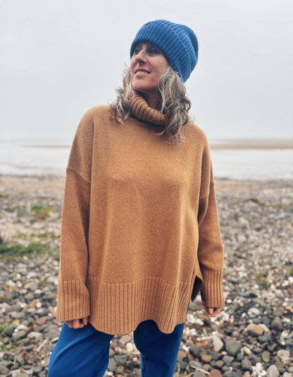 A woman wearing an oversized camel-colored knit turtleneck sweater, a blue beanie, and blue pants, standing on a pebble beach with water in the background.
