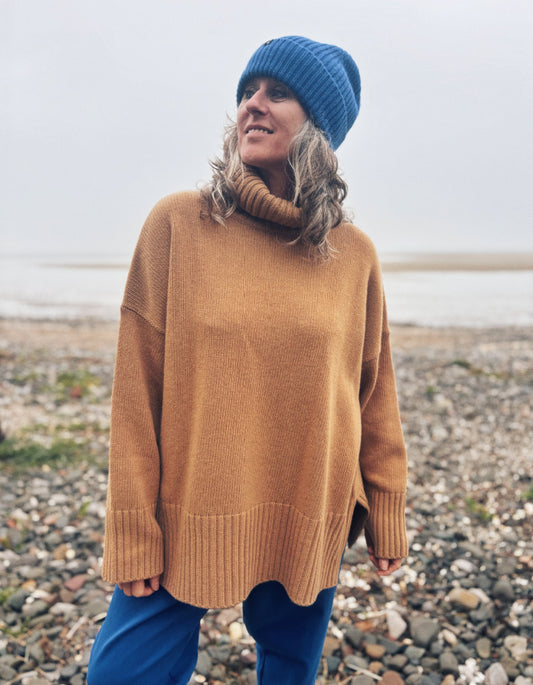 A woman wearing an oversized camel-colored knit turtleneck sweater, a blue beanie, and blue pants, standing on a pebble beach with water in the background.