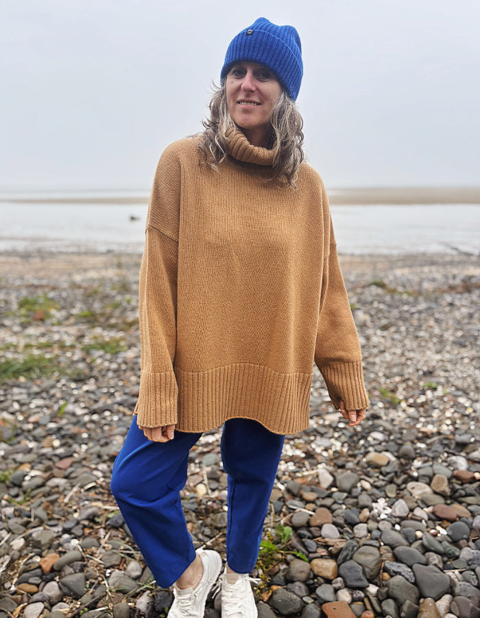A woman standing on a pebble beach wearing an oversized tan knit turtleneck sweater, bright blue pants, a blue beanie, and white sneakers.