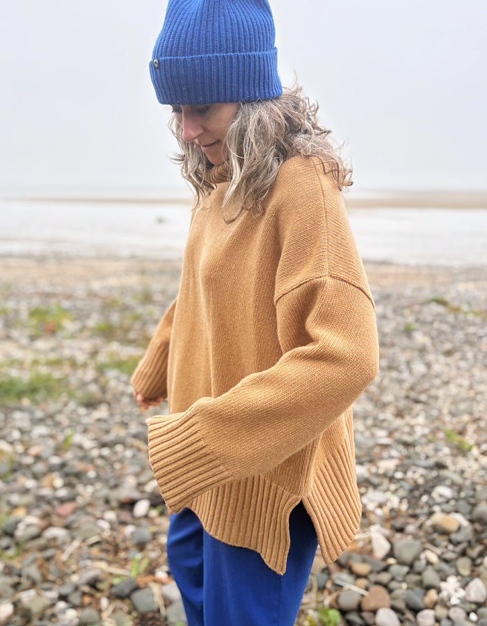 A person standing on a pebble beach wearing an oversized camel-colored knit sweater, a blue beanie, and blue pants.
