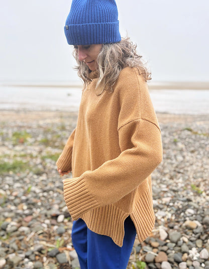 A person standing on a pebble beach wearing an oversized camel-colored knit sweater, a blue beanie, and blue pants.