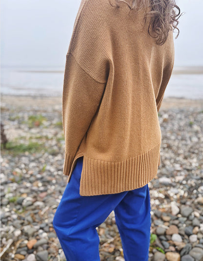A person standing on a pebble beach wearing an oversized tan knit sweater and bright blue pants, photographed from the side and back.