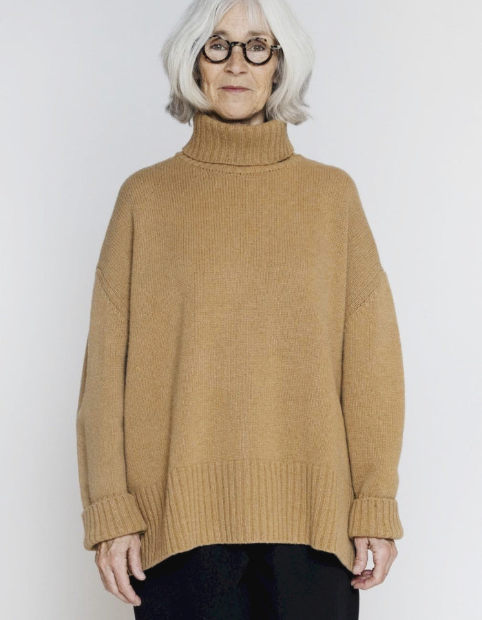 An older woman with short white hair and glasses wearing a camel-colored ribbed turtleneck sweater, standing against a light gray studio background.