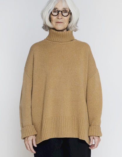 An older woman with short white hair and glasses wearing a camel-colored ribbed turtleneck sweater, standing against a light gray studio background.