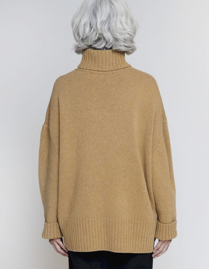 A person shown from the back wearing an oversized tan knit turtleneck sweater against a plain light gray studio background.