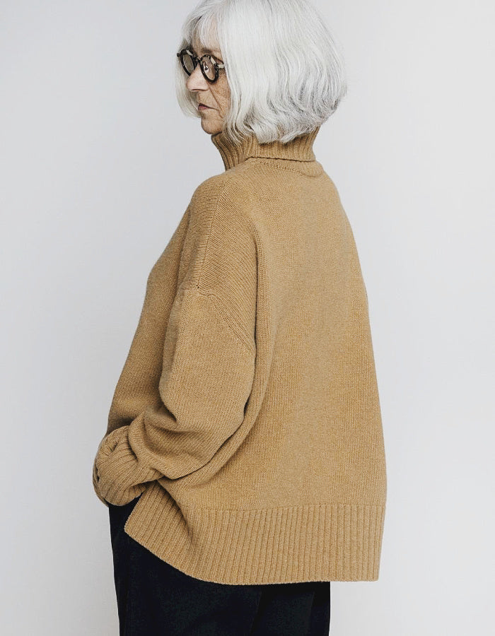 An older woman with short white hair and glasses, shown from a side-back angle wearing a camel-colored knit turtleneck sweater and dark pants against a light gray studio background.