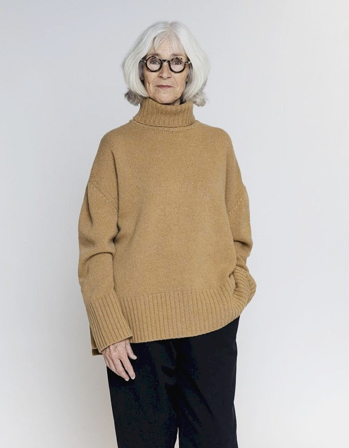 An older woman with short white hair and glasses wearing a camel-colored knit turtleneck sweater and black pants, standing against a light gray studio background.