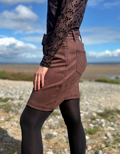 A person outdoors wearing a brown mini skirt with a side pocket, black tights, and a black patterned long-sleeve top, standing on a rocky beach with water and sky in the background.