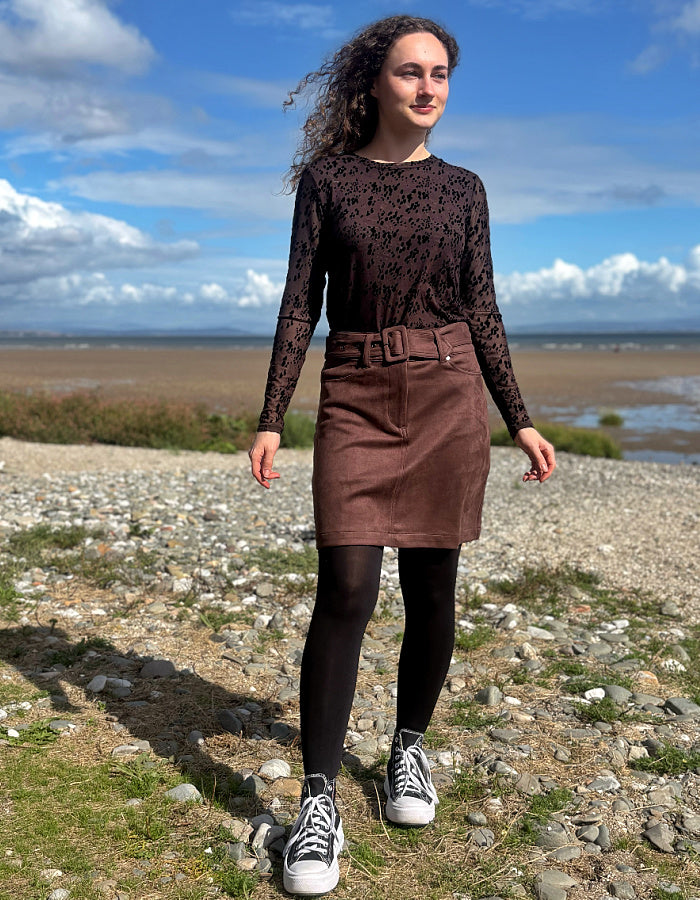 A woman stands on a rocky beach wearing a dark brown lace long-sleeve top, a brown corduroy skirt with a matching belt, black tights, and white canvas sneakers under a blue sky.