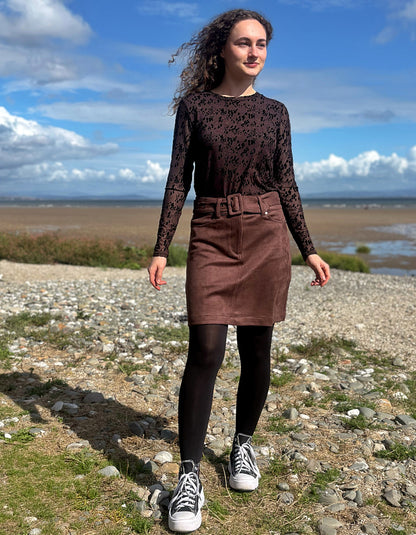 A woman stands on a rocky beach wearing a dark brown lace long-sleeve top, a brown corduroy skirt with a matching belt, black tights, and white canvas sneakers under a blue sky.