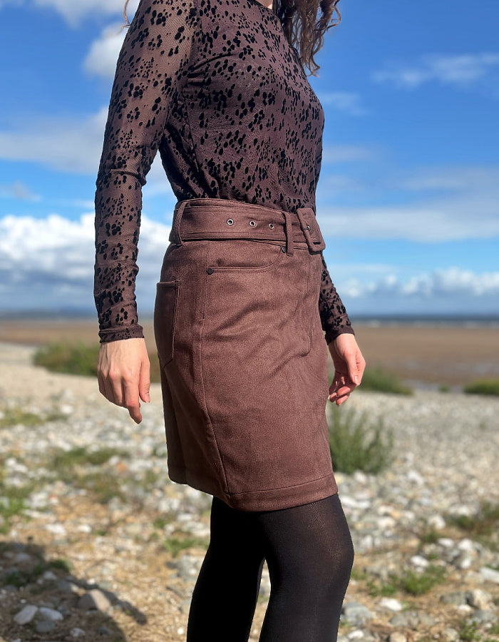 A person outdoors wearing a brown lace long-sleeve top and a brown suede-like mini skirt with a matching belt, paired with black tights. The background shows a rocky shoreline under a blue sky with clouds.