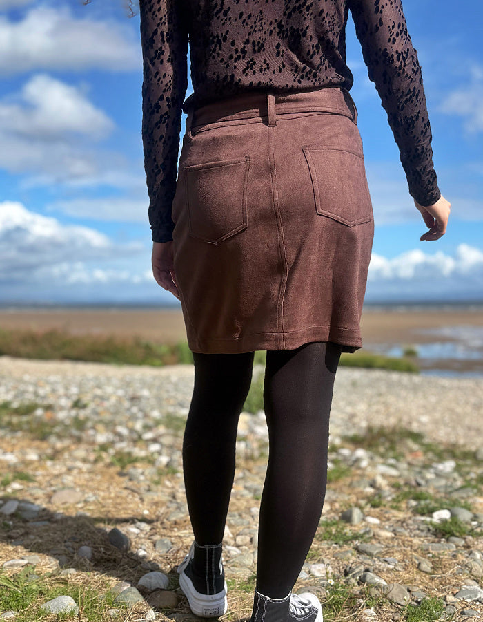 A person standing outdoors on a rocky beach or shoreline, shown from the back. The visible product appears to be a brown suede or faux-suede mini skirt with back patch pockets, worn over black tights and a dark lace long-sleeve top.