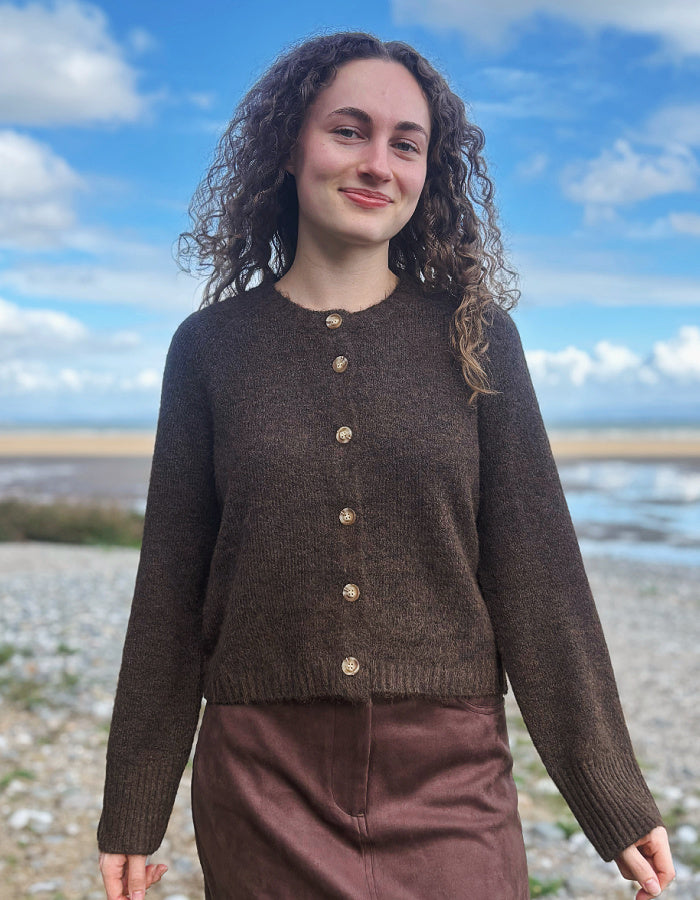 A woman with curly brown hair wearing a dark brown cropped knit cardigan with gold buttons and a brown skirt, standing outdoors on a pebble beach with water and a blue sky in the background.
