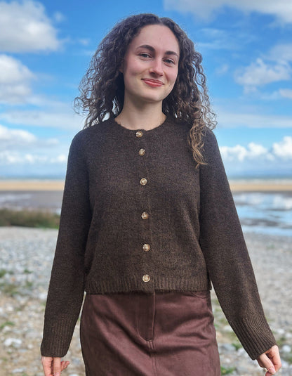 A woman with curly brown hair wearing a dark brown cropped knit cardigan with gold buttons and a brown skirt, standing outdoors on a pebble beach with water and a blue sky in the background.