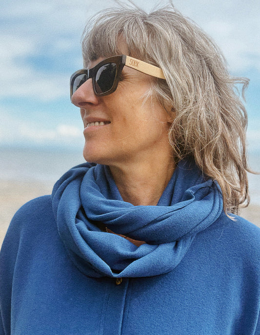 A woman outdoors wearing dark sunglasses with a light wood temple and a blue knit cowl scarf, dressed in a blue top against a soft-focus beach background.