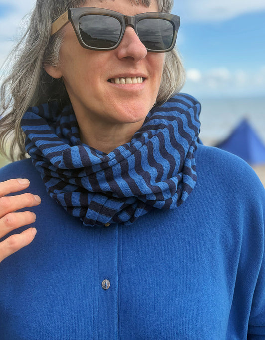 A close-up lifestyle shot of a person outdoors wearing a blue and black wavy striped infinity scarf, a blue button-up shirt, and sunglasses, with a blurred beach background.