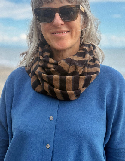 A woman outdoors wearing a brown and tan striped infinity scarf, a blue button-up cardigan, and sunglasses, with a blurred beach and sky background.