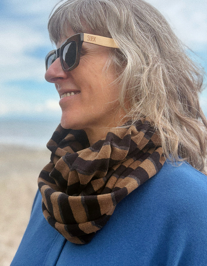 A woman outdoors wearing a brown and tan striped infinity scarf and wooden sunglasses with the brand name 'sox' visible on the temple.