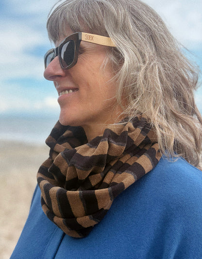 A woman outdoors wearing a brown and tan striped infinity scarf and wooden sunglasses with the brand name 'sox' visible on the temple.