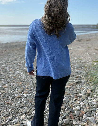 Person wearing a blue sweater standing on a pebbly beach with water and sky in the background