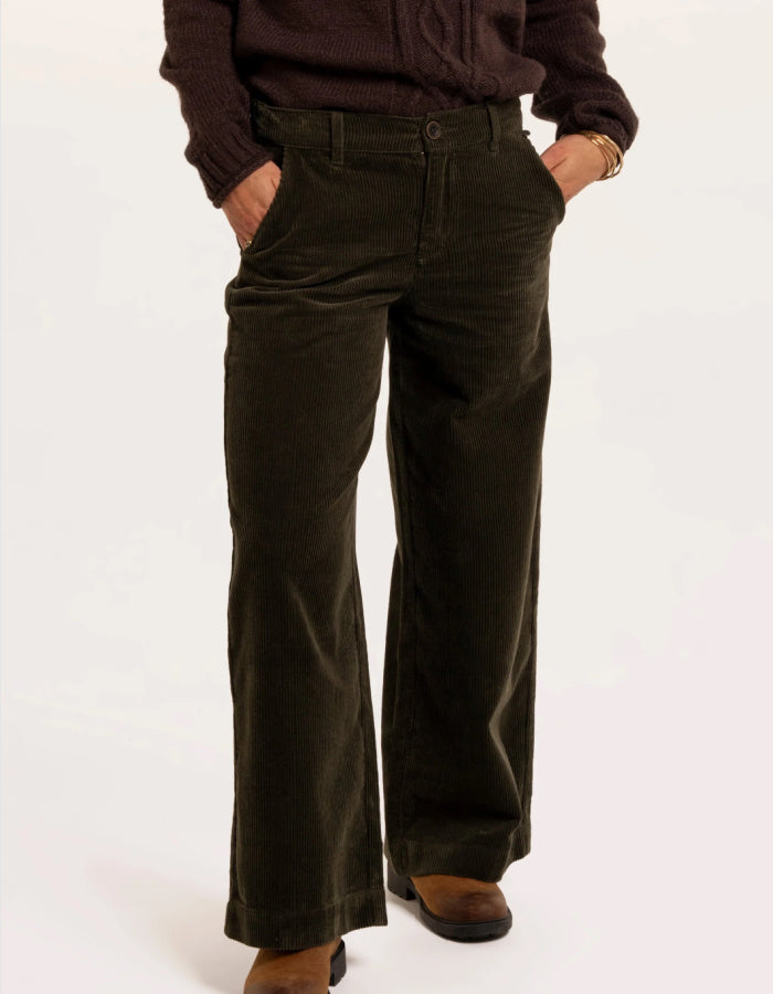 A person wearing dark olive green wide-leg corduroy pants with a dark brown sweater and brown boots, standing against a plain white studio background.