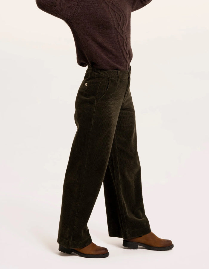 A person shown from the waist down wearing dark olive corduroy trousers, a dark brown sweater, and brown leather shoes against a plain white studio background.