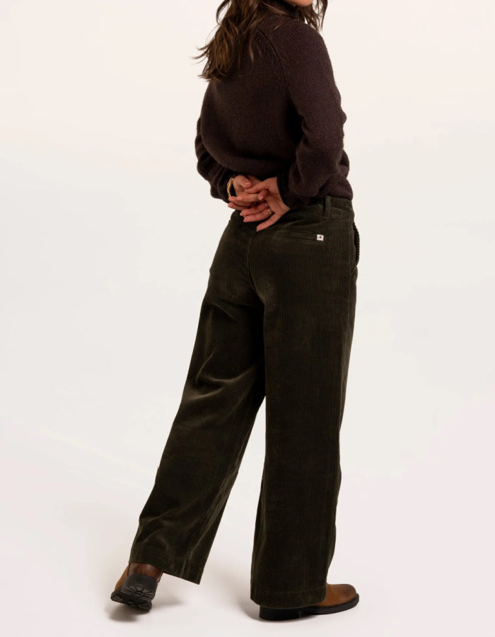 A model shown from the back wearing dark olive green wide-leg corduroy pants, a dark brown knit sweater, and brown leather shoes against a plain white studio background.