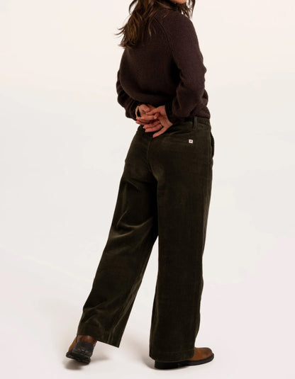 A model shown from the back wearing dark olive green wide-leg corduroy pants, a dark brown knit sweater, and brown leather shoes against a plain white studio background.