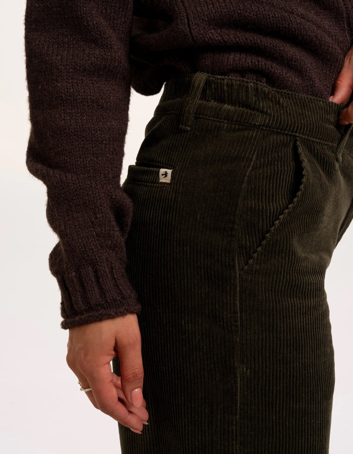 Close-up of a person wearing dark olive corduroy trousers and a dark brown ribbed knit sweater, showing the side pocket with a small white brand tab.