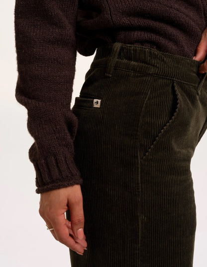 Close-up of a person wearing dark olive corduroy trousers and a dark brown ribbed knit sweater, showing the side pocket with a small white brand tab.