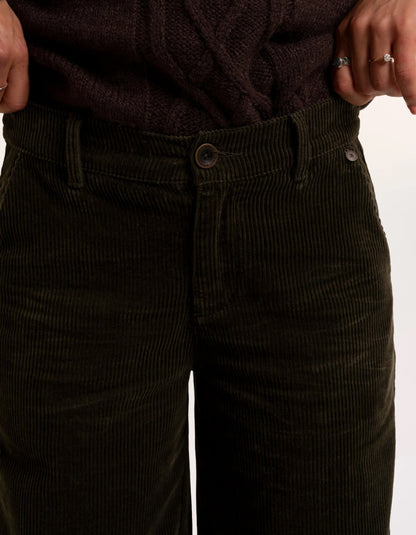 Close-up of a person wearing dark olive corduroy pants with a ribbed dark brown sweater tucked in, showing hands and rings against a plain light background.