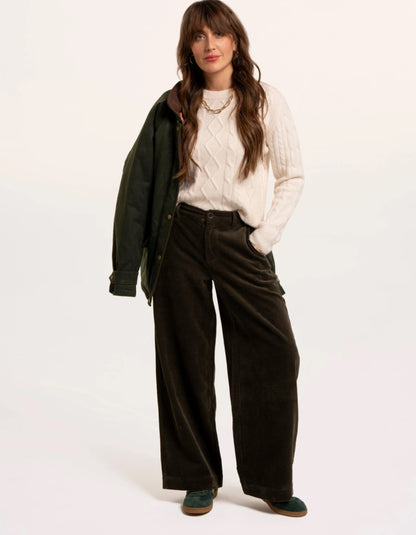 A woman modeling a white cable-knit sweater, dark brown wide-leg corduroy trousers, a dark green jacket draped over her shoulder, and green shoes, set against a plain off-white studio background.