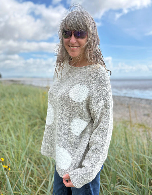 Person wearing a patterned boucle sweater standing in a grassy field with a blue sky and water in the background