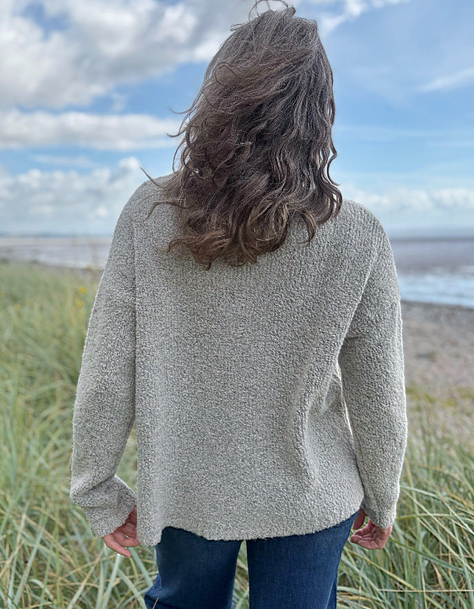 Person wearing a patterned boucle sweater standing in a grassy field with a blue sky and water in the background