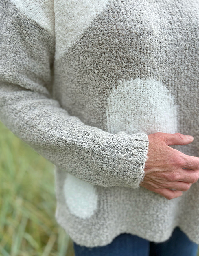 Person wearing a patterned boucle sweater standing in a grassy field with a blue sky and water in the background