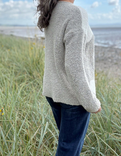 Person wearing a patterned boucle sweater standing in a grassy field with a blue sky and water in the background