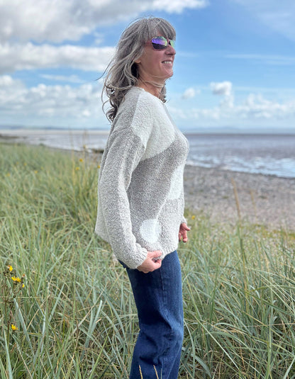 Person wearing a patterned boucle sweater standing in a grassy field with a blue sky and water in the background