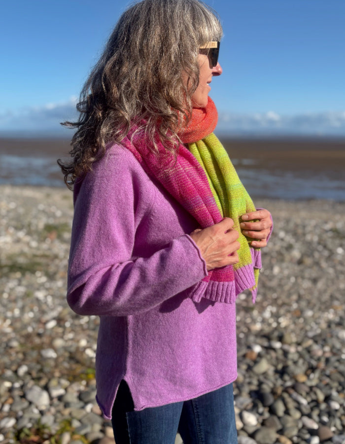 A woman standing on a rocky beach wearing a lavender-purple knit sweater, a bright pink and lime green gradient scarf, and blue jeans.