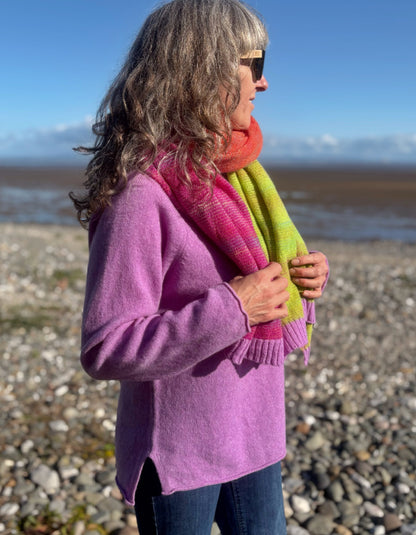 A woman standing on a rocky beach wearing a lavender-purple knit sweater, a bright pink and lime green gradient scarf, and blue jeans.