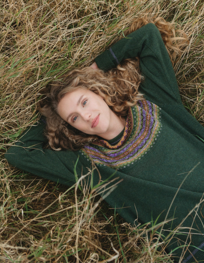 A woman with curly light brown hair lies in dry grass wearing a dark green knit sweater with a colorful patterned yoke.