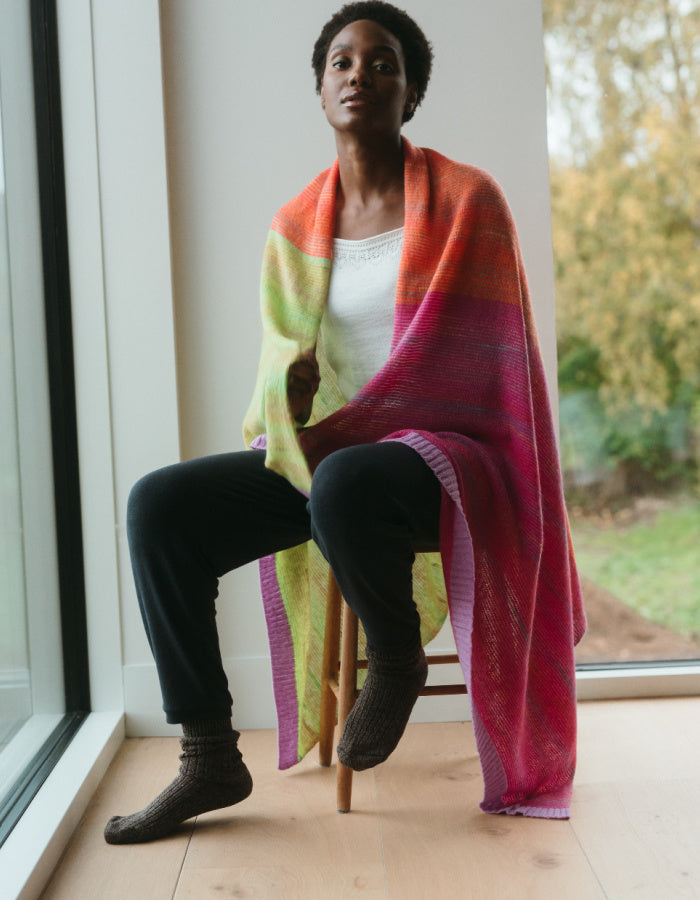 A woman sits on a wooden stool in a bright room by large windows, wrapped in a large knitted shawl with a gradient of orange, yellow, green, pink, and purple.