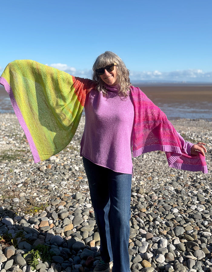 A woman standing on a rocky beach wearing a bright pink and lime green knit poncho or shawl over a lavender top and blue jeans, with her arms outstretched to show the full color of the garment.
