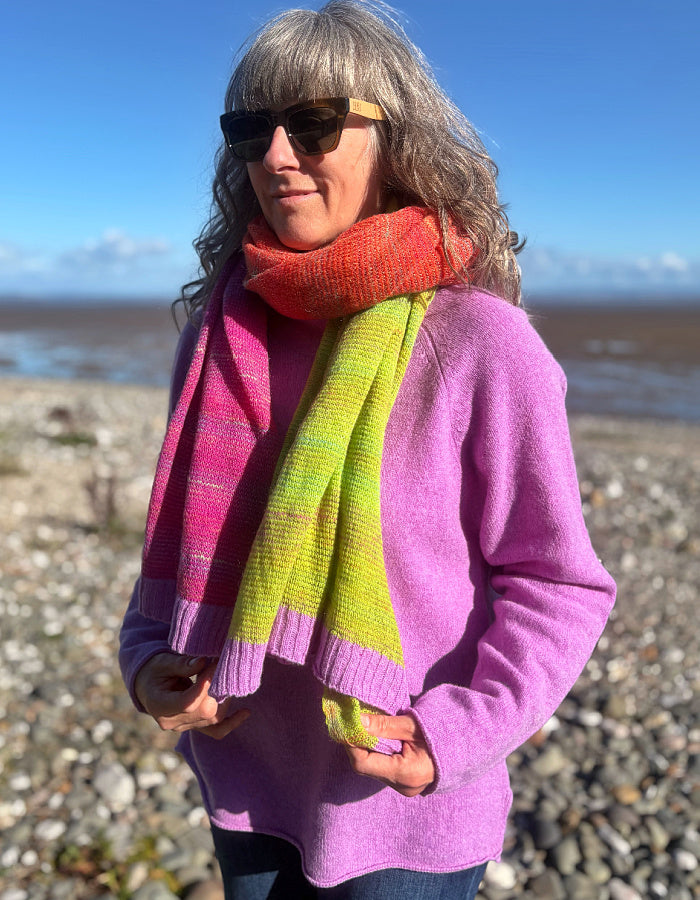 A woman standing on a pebble beach wearing a bright pink and lime green striped knit sweater and a matching striped knit scarf, with sunglasses and a blue sky background.
