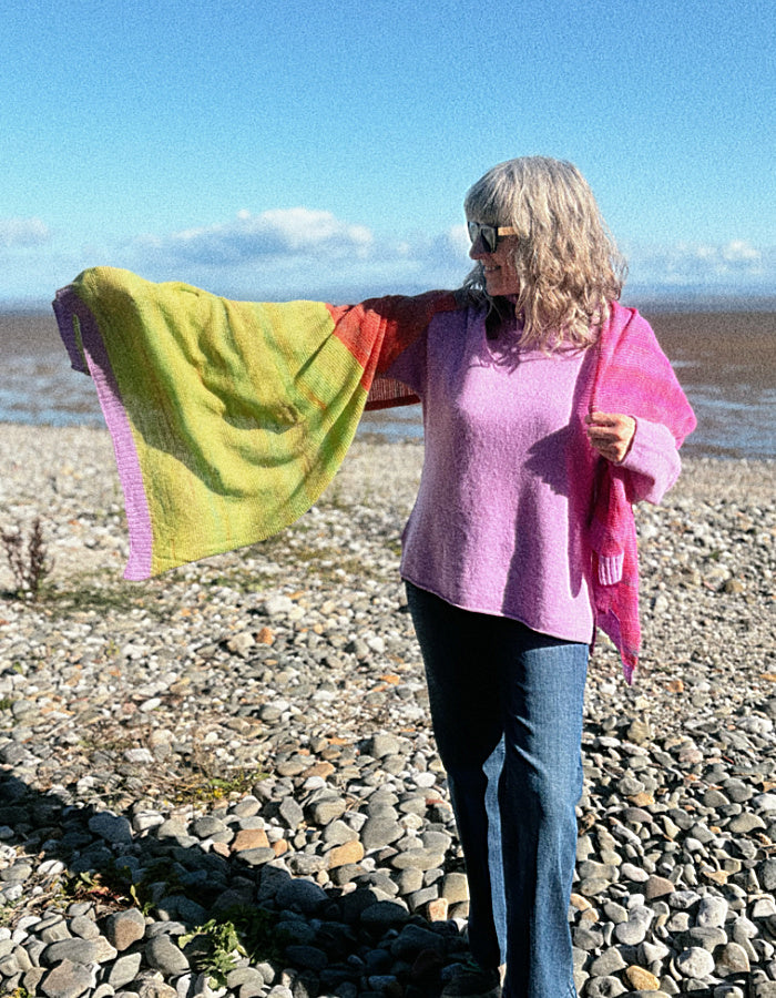 A woman standing on a pebble beach wearing a pink sweater and blue jeans, holding up a bright multicolored knit shawl or wrap with yellow, green, orange, and pink sections.