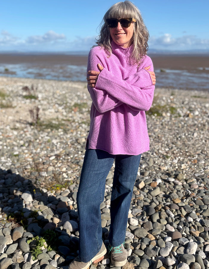 A woman standing on a rocky beach wearing a fuzzy light purple fleece turtleneck sweater, blue jeans, and sunglasses.