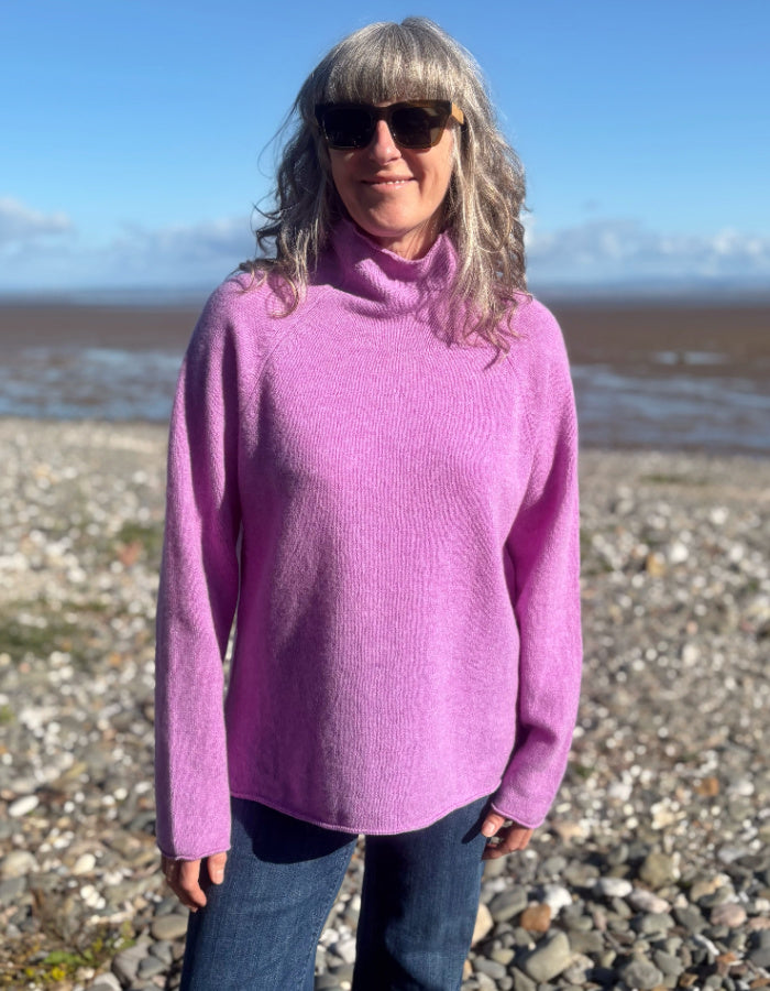 A woman with gray hair and sunglasses wearing a bright pink turtleneck sweater and blue jeans, standing on a rocky beach with water and a blue sky in the background.