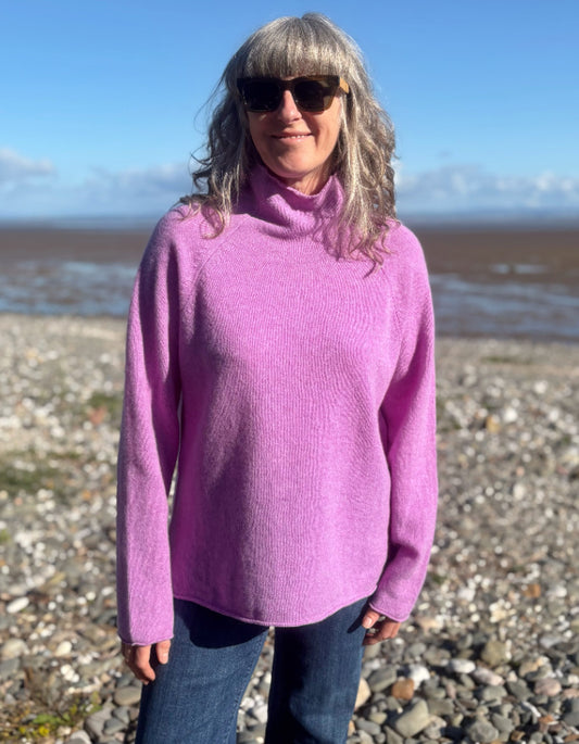 A woman with gray hair and sunglasses wearing a bright pink turtleneck sweater and blue jeans, standing on a rocky beach with water and a blue sky in the background.