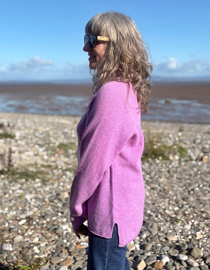 A woman with long gray hair and sunglasses stands in profile on a pebble beach, wearing a fuzzy lavender-pink knit cardigan and dark blue jeans, with water and a blue sky in the background.